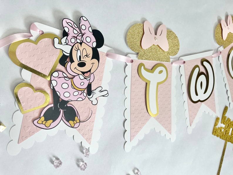 K&ouml;nnte beinhalten: Rosa-goldener Minnie Mouse-Banner mit dem Wort "Zwei" in goldenen Buchstaben. Der Banner hat einen rosa Hintergrund mit wei&szlig;en gewellten R&auml;ndern und eine Minnie Mouse-Figur.