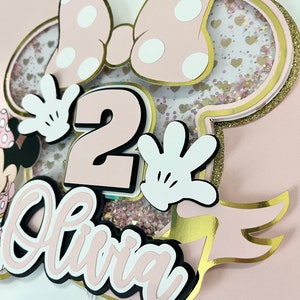 K&ouml;nnte beinhalten: Rosa-goldener Minnie Mouse-Tortenaufsatz mit einer wei&szlig;en Schleife, Glitzer und der Zahl 2. Der Name Olivia ist in rosa und schwarzen Buchstaben geschrieben.