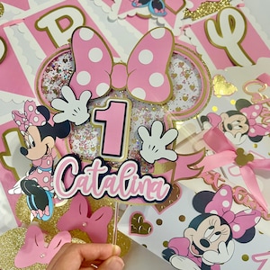 K&ouml;nnte beinhalten: Rosa und goldene Minnie Mouse Partydekorationen mit einem Tortendekor mit einer rosa Schleife, einer Nummer eins und dem Namen "Catalina".