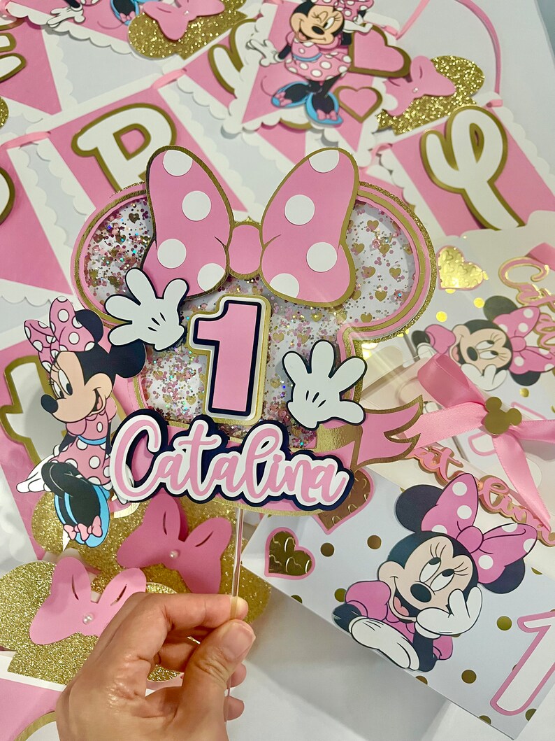 K&ouml;nnte beinhalten: Rosa und goldene Minnie Mouse Partydekorationen mit einem Tortendekor mit einer rosa Schleife, einer Nummer eins und dem Namen "Catalina".