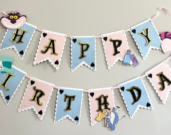 May include: A pink and blue banner with the words "Happy Birthday" in gold letters. The banner is decorated with hearts, a Cheshire Cat, the Mad Hatter, a bottle of potion, a playing card, and a pocket watch.