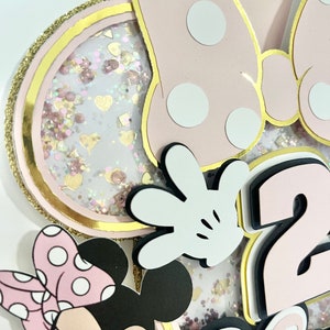 K&ouml;nnte beinhalten: Rosa-schwarzer Minnie Mouse-Kuchentopper mit einer gro&szlig;en rosa Schleife mit wei&szlig;en Punkten, einem wei&szlig;en Handschuh, einer rosa Zahl 2 und dem Wort "Zwei" in rosa. Der Topper ist mit rosa und goldenen Glitzer verziert.