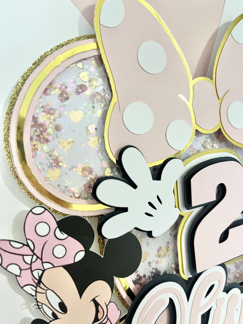 K&ouml;nnte beinhalten: Rosa-schwarzer Minnie Mouse-Kuchentopper mit einer gro&szlig;en rosa Schleife mit wei&szlig;en Punkten, einem wei&szlig;en Handschuh, einer rosa Zahl 2 und dem Wort "Zwei" in rosa. Der Topper ist mit rosa und goldenen Glitzer verziert.