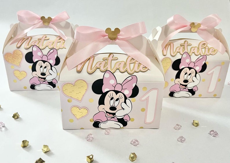 K&ouml;nnte beinhalten: Drei rosa und goldene Geschenkboxen mit Minnie Mouse-Motiven. Jede Box hat einen rosa Schleifen-Griff und ein goldenes Herz-Muster. Die Boxen sind mit einem Minnie Mouse-Charakter, einer goldenen Zahl eins und dem Namen "Natalie" verziert.