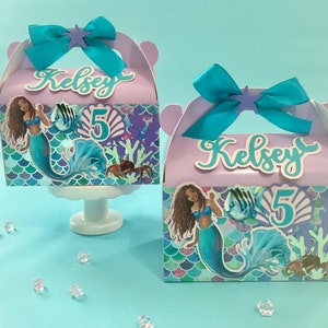 May include: Two purple and blue mermaid-themed party favor boxes with a mermaid, fish, and seashells design. The boxes have a handle and say "Kelsey 5" on the front.