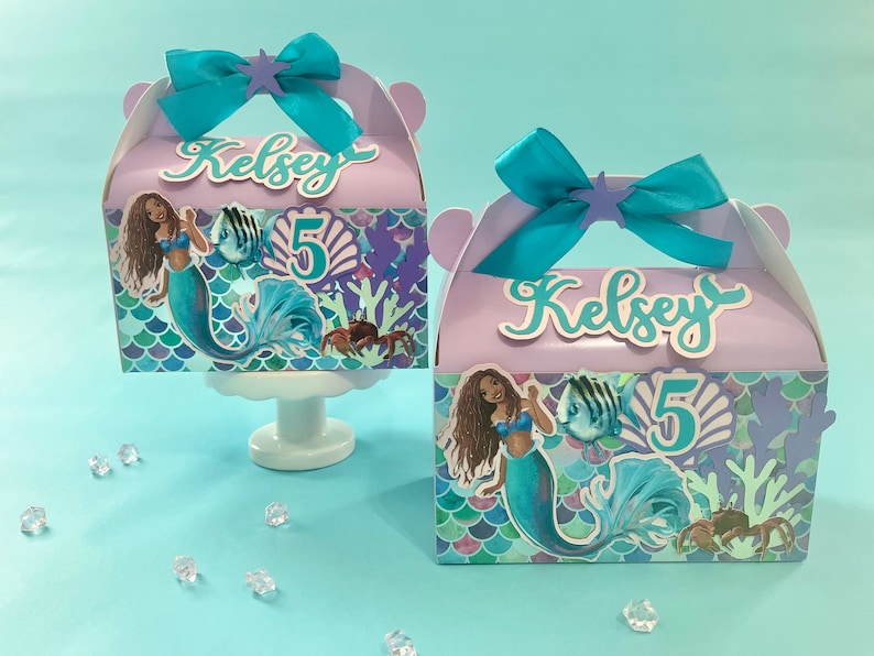 May include: Two purple and blue mermaid-themed party favor boxes with a mermaid, fish, and seashells design. The boxes have a handle and say "Kelsey 5" on the front.