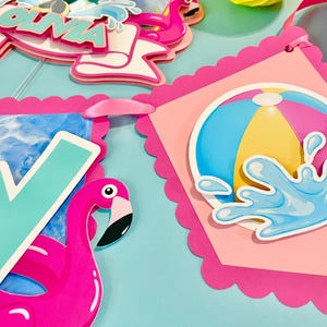 May include: A decorative party banner featuring scalloped pink edges, a large teal letter W, a bright pink flamingo, and a circular graphic of a colorful beach ball with a blue water splash effect. The design includes layered paper cutouts.