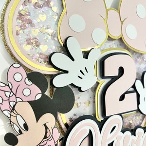 K&ouml;nnte beinhalten: Rosa-schwarzer Minnie Mouse-Kuchentopper mit einer gro&szlig;en rosa Zahl 2, dem Wort "Alice" in rosa Schrift und einer kleinen Minnie Mouse-Figur. Der Topper hat einen glitzernden Hintergrund.