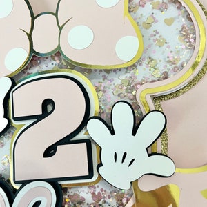 K&ouml;nnte beinhalten: Rosa und goldene Minnie Mouse-Geburtstagsdekorationen mit einer Nummer 2, einem wei&szlig;en Handschuh und Glitzer.