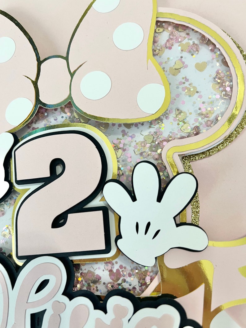 K&ouml;nnte beinhalten: Rosa und goldene Minnie Mouse-Geburtstagsdekorationen mit einer Nummer 2, einem wei&szlig;en Handschuh und Glitzer.