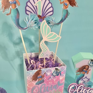 May include: A mermaid-themed centerpiece for a first birthday party. The centerpiece includes a purple and blue seashell with the number one, a mermaid tail, and two mermaid figures. The centerpiece is in a square box with a mermaid design and the name "Olivia" written on it.