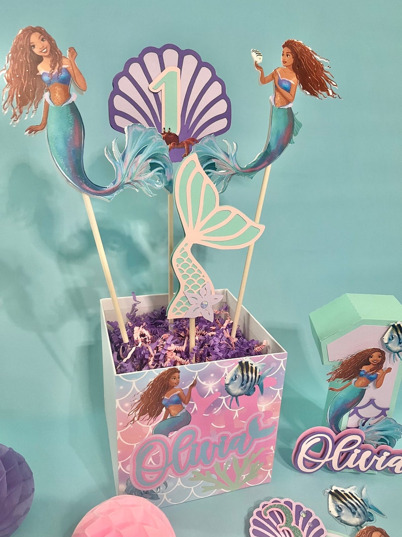 May include: A mermaid-themed centerpiece for a first birthday party. The centerpiece includes a purple and blue seashell with the number one, a mermaid tail, and two mermaid figures. The centerpiece is in a square box with a mermaid design and the name "Olivia" written on it.