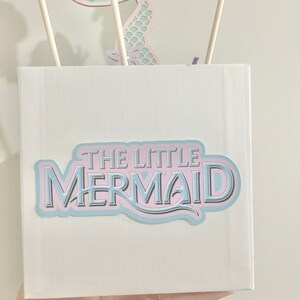 May include: A white box with a blue and pink sticker that says "THE LITTLE MERMAID". The box is decorated with a cutout of a mermaid tail.
