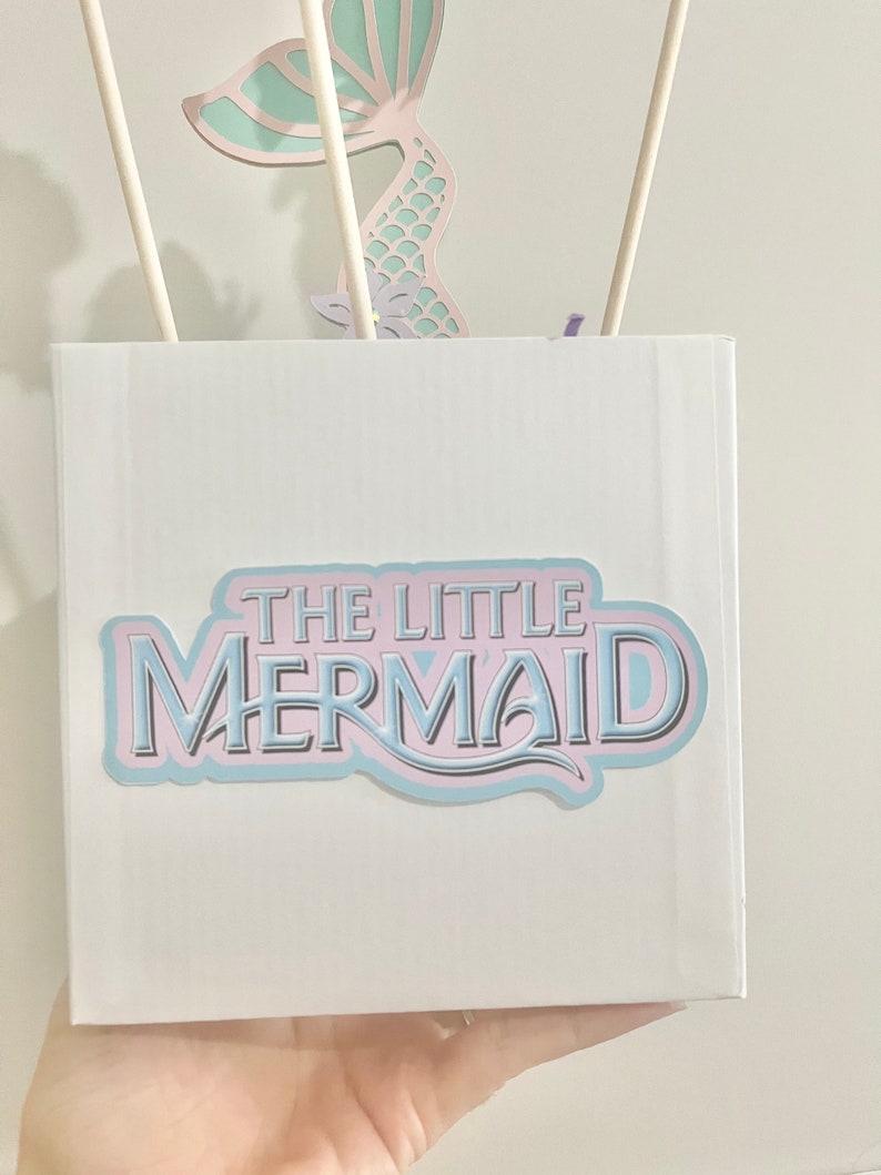 May include: A white box with a blue and pink sticker that says "THE LITTLE MERMAID". The box is decorated with a cutout of a mermaid tail.