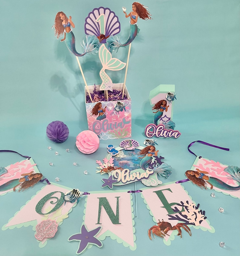 May include: A mermaid-themed first birthday party centerpiece with a purple and pink color scheme. The centerpiece includes a cake topper, a banner, and a centerpiece box with a mermaid figurine and the name "Olivia".