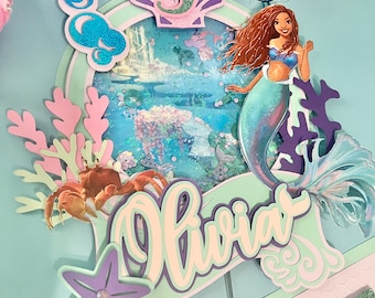 May include: A mermaid-themed cake topper with a purple seashell, a mermaid with brown hair, a crab, a starfish, and the name "Olivia" in a green and purple font.