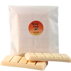 May include: Two white chocolate bars with a Stay-Cay sticker on a white paper bag. The sticker says "Stay-Cay" in a circle with a wave design and the text "please do not disturb, Pineapple, Mango, & Couch Surfing".