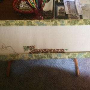 May include: A close-up of a cross-stitch project in progress, featuring a giraffe with brown and tan spots, set in a wooden embroidery hoop with a green and blue floral patterned fabric.