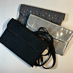 May include: Three different fabric clutches, one black with a floral pattern, one silver with a sparkly finish, and one gray with a pattern of arrows. The black clutch has a black strap.