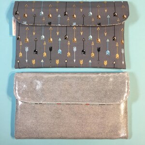 May include: Two gray clutches. The top clutch is made of fabric with a pattern of gold, black, and blue arrows. The bottom clutch is made of a shimmery, textured material.