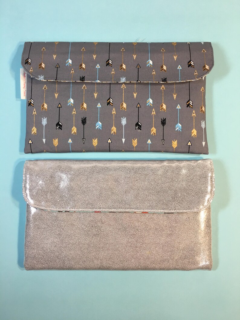 May include: Two gray clutches. The top clutch is made of fabric with a pattern of gold, black, and blue arrows. The bottom clutch is made of a shimmery, textured material.