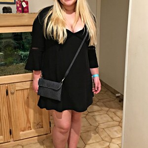 May include: A woman wearing a black dress with a plunging neckline and a black crossbody bag. She is wearing black ankle boots and has long blonde hair.