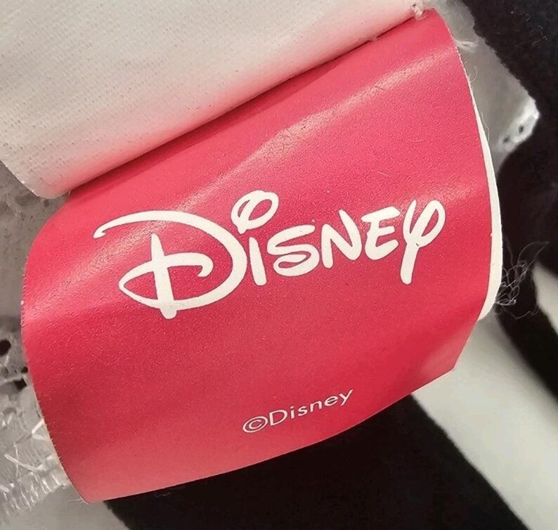 Peut inclure: Une &eacute;tiquette rouge Disney avec le logo Disney en blanc. L'&eacute;tiquette est attach&eacute;e &agrave; un article noir et blanc. Le logo Disney est une &eacute;criture stylis&eacute;e avec une ic&ocirc;ne de ch&acirc;teau au-dessus du mot Disney. L'&eacute;tiquette comprend &eacute;galement le symbole du droit d'auteur et le mot "Disney".