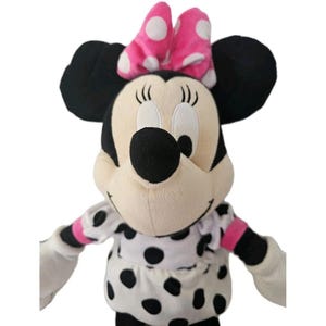 Pode incluir: Um peluche de uma personagem rato com grandes orelhas pretas, rosto cor de creme e nariz preto. A personagem usa um la&ccedil;o cor-de-rosa brilhante com bolinhas brancas e um vestido branco com bolinhas pretas e detalhes cor-de-rosa nas mangas.