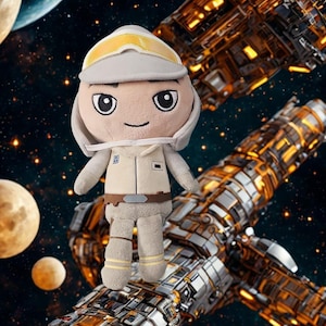 Puede incluir: Un muñeco de peluche de un personaje con traje de vuelo color canela, una gorra a juego con gafas amarillas y un cinturón marrón. El muñeco tiene grandes ojos negros bordados, una boca sencilla y una tez de color canela claro.
