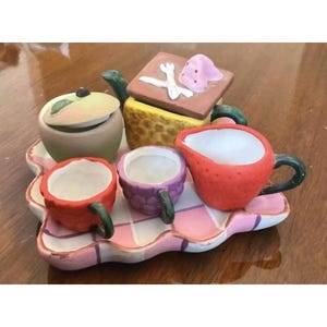 May include: A miniature ceramic tea set on a pink and white checkered tray. The set includes a green lidded sugar bowl, a yellow square teapot with a pink lid, a red strawberry-shaped creamer, and two small cups in red and purple.