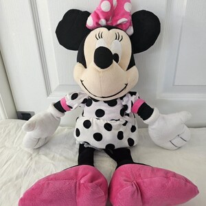 Peut inclure: Poup&eacute;e en peluche Minnie Mouse avec une t&ecirc;te et des oreilles noires, un n&oelig;ud rose &agrave; pois blancs et une robe blanche &agrave; pois noirs. La poup&eacute;e a des gants blancs et des chaussures roses.