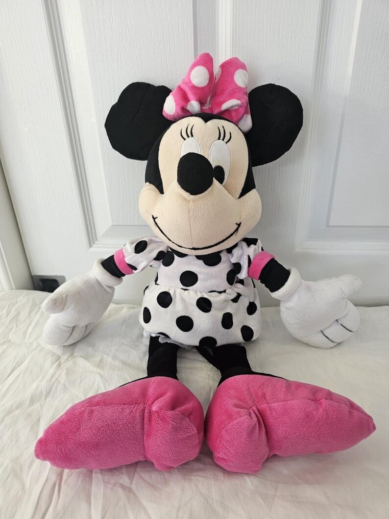 Peut inclure: Poup&eacute;e en peluche Minnie Mouse avec une t&ecirc;te et des oreilles noires, un n&oelig;ud rose &agrave; pois blancs et une robe blanche &agrave; pois noirs. La poup&eacute;e a des gants blancs et des chaussures roses.