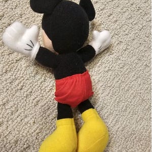 Puede incluir: Un mu&ntilde;eco de peluche de Mickey Mouse, con cabeza y orejas negras, guantes blancos, pantalones cortos rojos y zapatos amarillos. El mu&ntilde;eco est&aacute; sobre una superficie clara y texturizada.