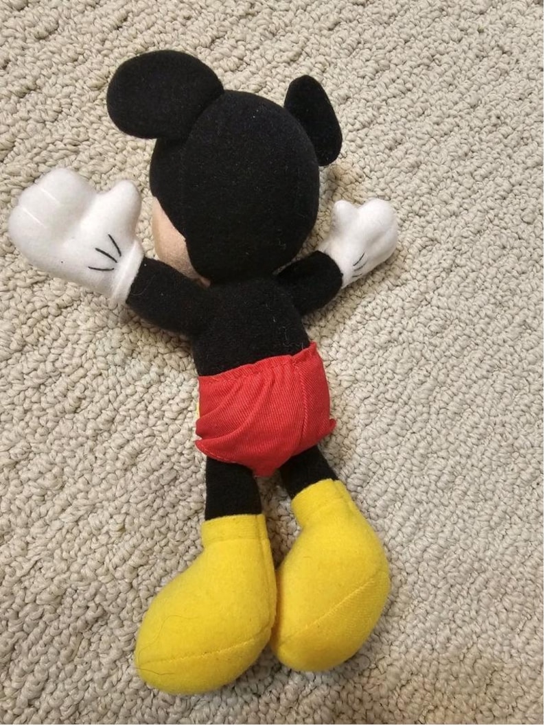 Puede incluir: Un mu&ntilde;eco de peluche de Mickey Mouse, con cabeza y orejas negras, guantes blancos, pantalones cortos rojos y zapatos amarillos. El mu&ntilde;eco est&aacute; sobre una superficie clara y texturizada.