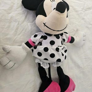 Peut inclure: Une poup&eacute;e en peluche Minnie Mouse. La poup&eacute;e a une t&ecirc;te noire avec de grandes oreilles, un visage blanc et un n&oelig;ud rose &agrave; pois blancs. Elle porte une robe blanche &agrave; pois noirs, des jambes noires et des chaussures roses.