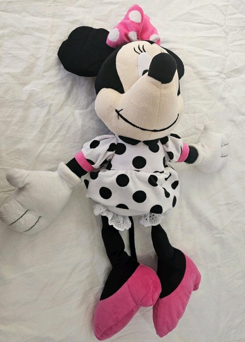 Peut inclure: Une poup&eacute;e en peluche Minnie Mouse. La poup&eacute;e a une t&ecirc;te noire avec de grandes oreilles, un visage blanc et un n&oelig;ud rose &agrave; pois blancs. Elle porte une robe blanche &agrave; pois noirs, des jambes noires et des chaussures roses.