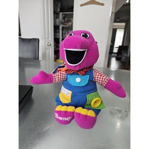 Op de afbeelding: Een paarse pluche dinosaurus met een rood-wit geblokt overhemd, een rood gestreepte vlinderdas en een blauwe tuinbroek. De tuinbroek heeft een turquoise zak met een witte knoop, een gele zak met een drukknoop en een groene zak met een knoop.