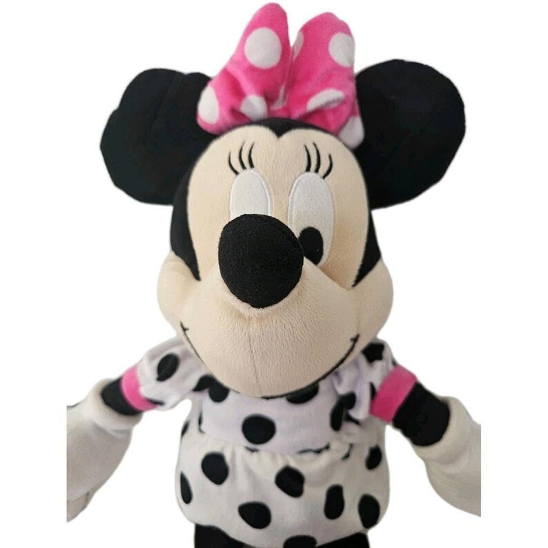Peut inclure: Une poup&eacute;e en peluche Minnie Mouse avec des oreilles noires, un n&oelig;ud rose &agrave; pois blancs et une robe &agrave; pois noirs et blancs. La poup&eacute;e a un nez noir, des yeux blancs et une expression souriante. Les bras sont blancs avec des accents roses.