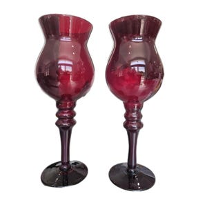 May include: A pair of tall, deep red glass goblets featuring a bulbous bowl shape, a slender stem with three distinct decorative rings, and a wide, circular base. The translucent glass reflects light, highlighting the rich crimson colour throughout.