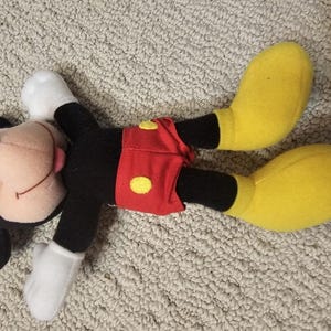 Puede incluir: Un peluche de Mickey Mouse acostado sobre una superficie texturizada. El juguete presenta un cuerpo negro, guantes blancos y zapatos amarillos grandes. Mickey lleva pantalones cortos rojos con botones amarillos y tiene una cara sonriente con una lengua rosa.