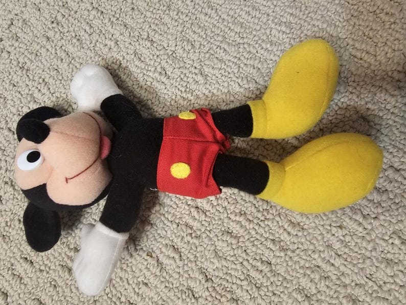 Puede incluir: Un peluche de Mickey Mouse acostado sobre una superficie texturizada. El juguete presenta un cuerpo negro, guantes blancos y zapatos amarillos grandes. Mickey lleva pantalones cortos rojos con botones amarillos y tiene una cara sonriente con una lengua rosa.