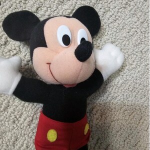 Puede incluir: Un peluche de Mickey Mouse con orejas negras, cuerpo negro y pantalones cortos rojos con botones amarillos. El juguete tiene guantes blancos y boca rosa. El fondo es una superficie clara y texturizada.