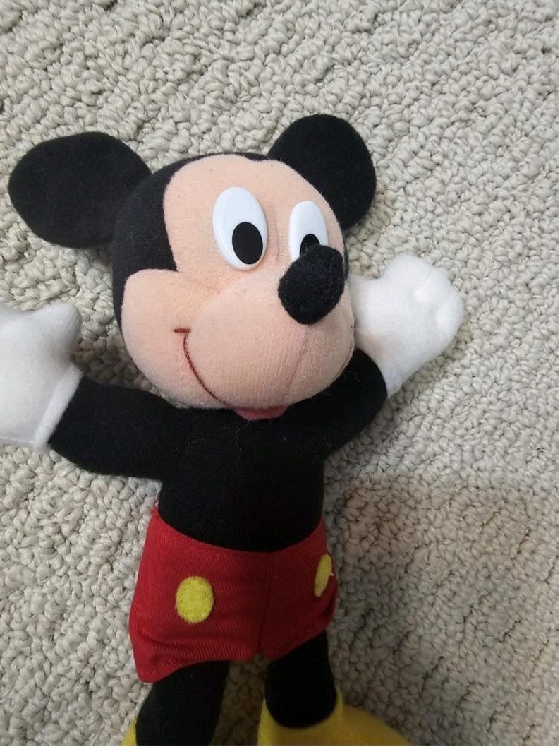 Puede incluir: Un peluche de Mickey Mouse con orejas negras, cuerpo negro y pantalones cortos rojos con botones amarillos. El juguete tiene guantes blancos y boca rosa. El fondo es una superficie clara y texturizada.