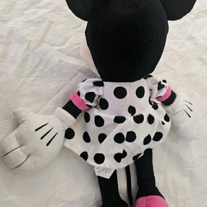 Pode incluir: Um brinquedo de pel&uacute;cia de um personagem de rato de desenho animado com grandes orelhas pretas, um la&ccedil;o rosa com bolinhas brancas e um vestido branco com bolinhas pretas. O personagem tem luvas brancas, membros pretos e sapatos rosa com etiqueta.