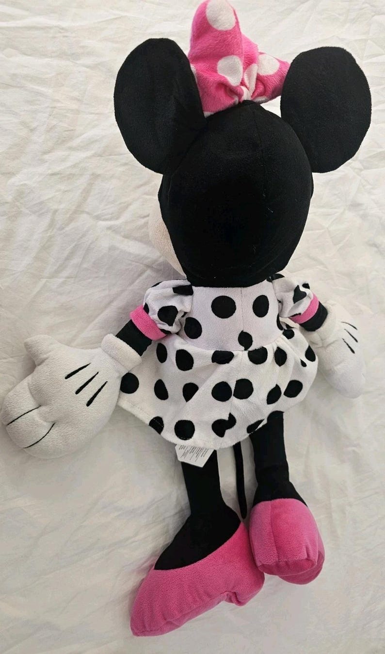 Pode incluir: Um brinquedo de pel&uacute;cia de um personagem de rato de desenho animado com grandes orelhas pretas, um la&ccedil;o rosa com bolinhas brancas e um vestido branco com bolinhas pretas. O personagem tem luvas brancas, membros pretos e sapatos rosa com etiqueta.