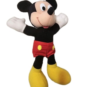 Puede incluir: Un peluche de Mickey Mouse con orejas negras, cuerpo negro y cara beige. Mickey lleva pantalones cortos rojos con botones amarillos, guantes blancos y zapatos amarillos. Los brazos del juguete est&aacute;n extendidos.