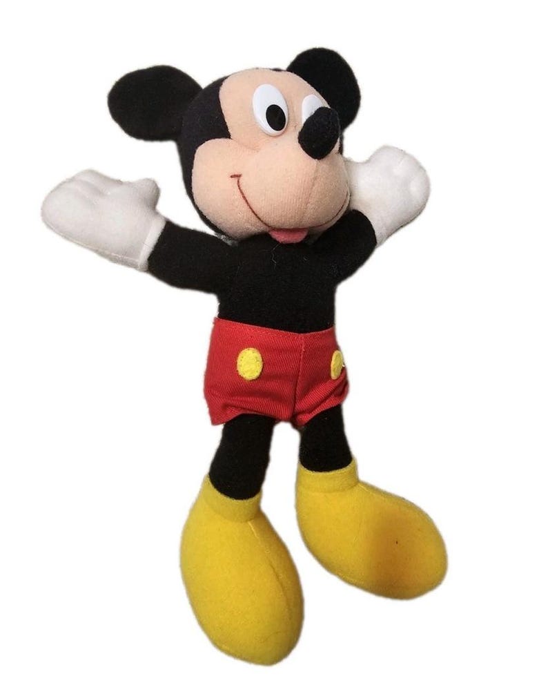 Puede incluir: Un peluche de Mickey Mouse con orejas negras, cuerpo negro y cara beige. Mickey lleva pantalones cortos rojos con botones amarillos, guantes blancos y zapatos amarillos. Los brazos del juguete est&aacute;n extendidos.