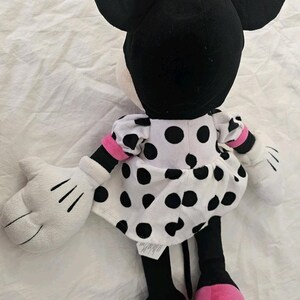 Peut inclure: Une poup&eacute;e en peluche de Minnie Mouse, vue de dos. La poup&eacute;e pr&eacute;sente de grandes oreilles noires, un n&oelig;ud rose &agrave; pois blancs et une robe blanche &agrave; pois noirs. Les mains de la poup&eacute;e sont blanches et les pieds roses.
