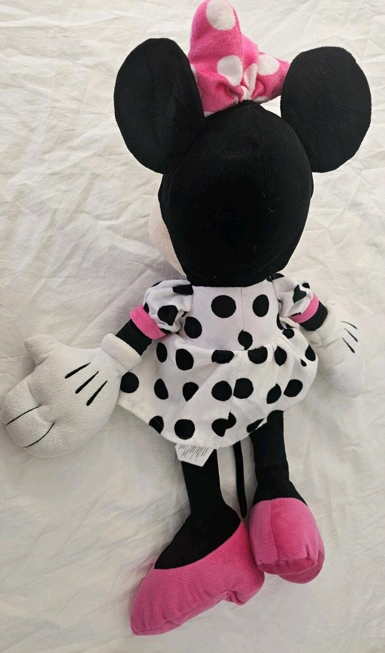 Peut inclure: Une poup&eacute;e en peluche de Minnie Mouse, vue de dos. La poup&eacute;e pr&eacute;sente de grandes oreilles noires, un n&oelig;ud rose &agrave; pois blancs et une robe blanche &agrave; pois noirs. Les mains de la poup&eacute;e sont blanches et les pieds roses.