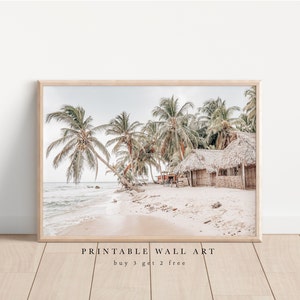 Puede incluir: Una impresión de arte de pared imprimible que presenta una escena de playa con palmeras y cabañas con techo de paja. La imagen está en una paleta de colores claros y aireados con una sensación suave y de ensueño. El texto "PRINTABLE WALL ART" y "buy 3 get 2 free" está en la parte inferior de la imagen.